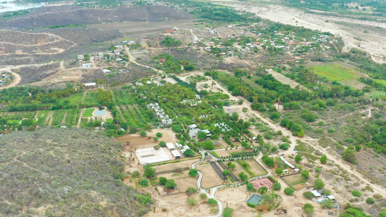 Opening aerial frame over La Playita Los Cabos with dry scrub hills, green fields, dirt tracks, small farm buildings, and no visible people.