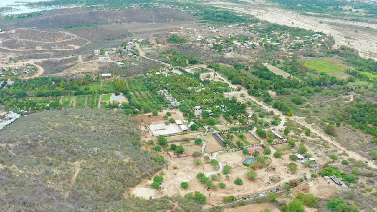 High aerial view over La Playita Los Cabos rural fields, pale dirt roads, scattered trees, dry hills, small structures, and no visible people at the start of the clip.