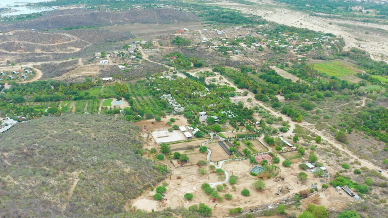 Wide aerial view of La Playita Los Cabos countryside with a pale road crossing between green fields, low dry hills with visible tracks, scattered structures, and no visible people.