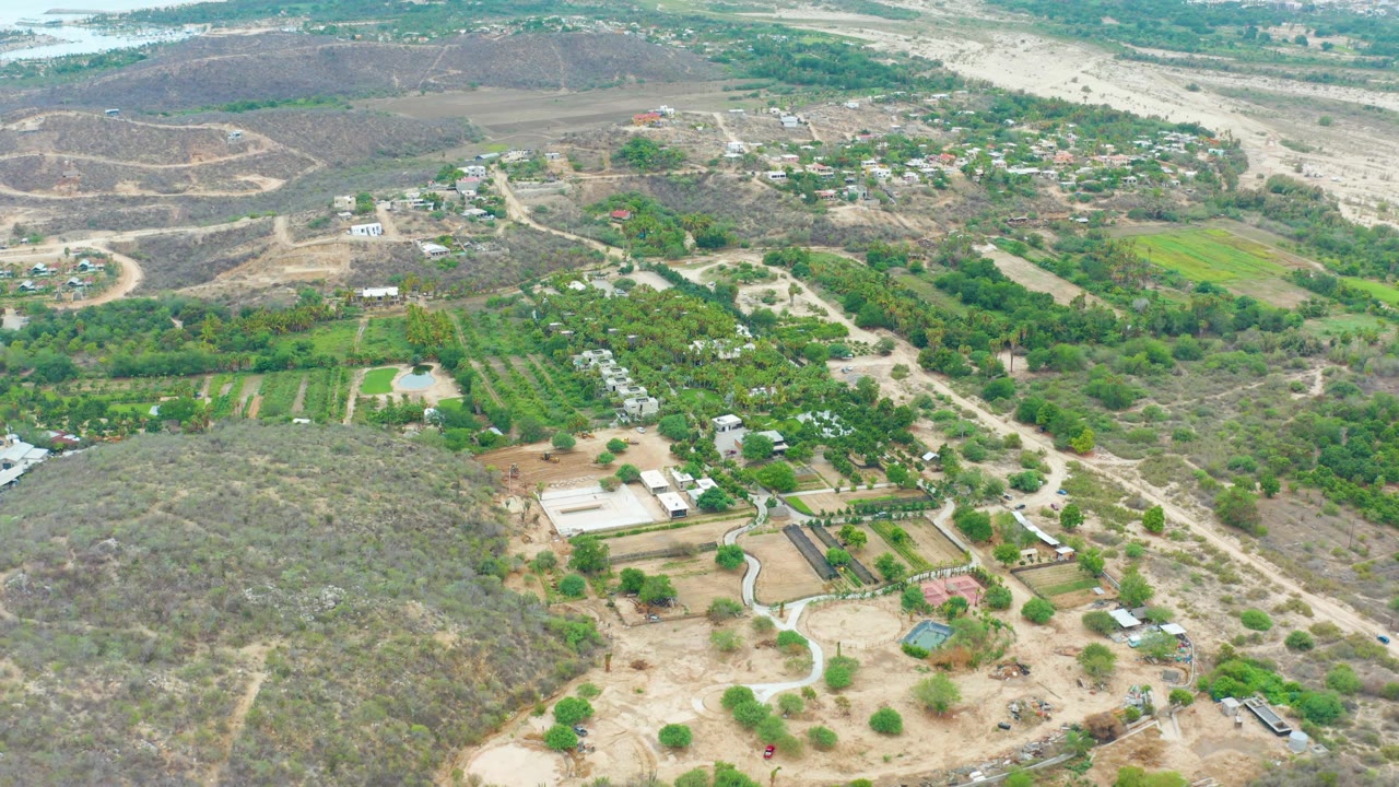 Aerial rural road scene with farm plots, trees, buildings, and dry terrain.