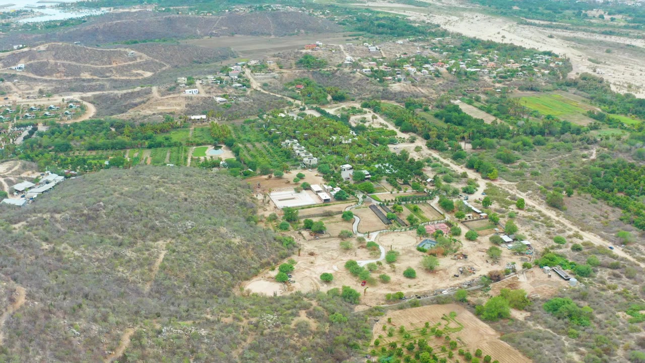 High aerial view over La Playita Los Cabos with green farm plots, brown scrubland, dirt roads, scattered buildings, and a pale river corridor in the background, with no visible people.