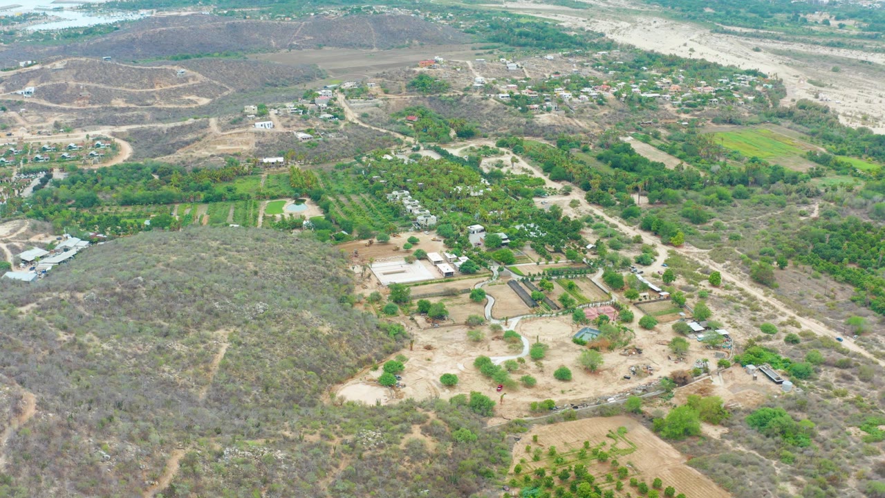 Wide overhead view of La Playita Los Cabos farmland with green vegetation blocks, dusty roads, brown hillsides, scattered buildings, and the light-toned waterway corridor across the top of frame, no visible people.