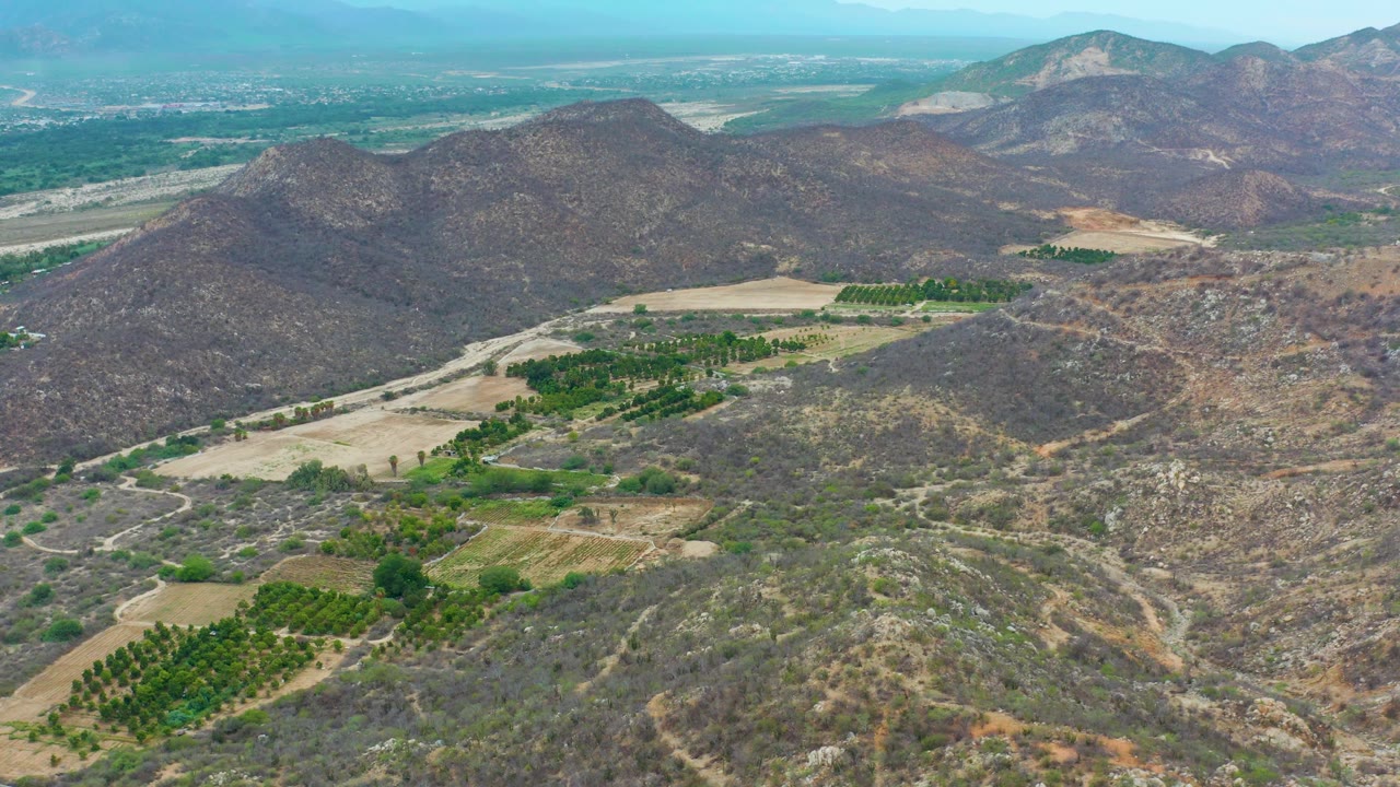 Wide aerial frame from La Playita Los Cabos, Mexico, showing arid mountain slopes, green cultivated patches, pale open fields, a distant urban edge near the horizon, and no visible people.