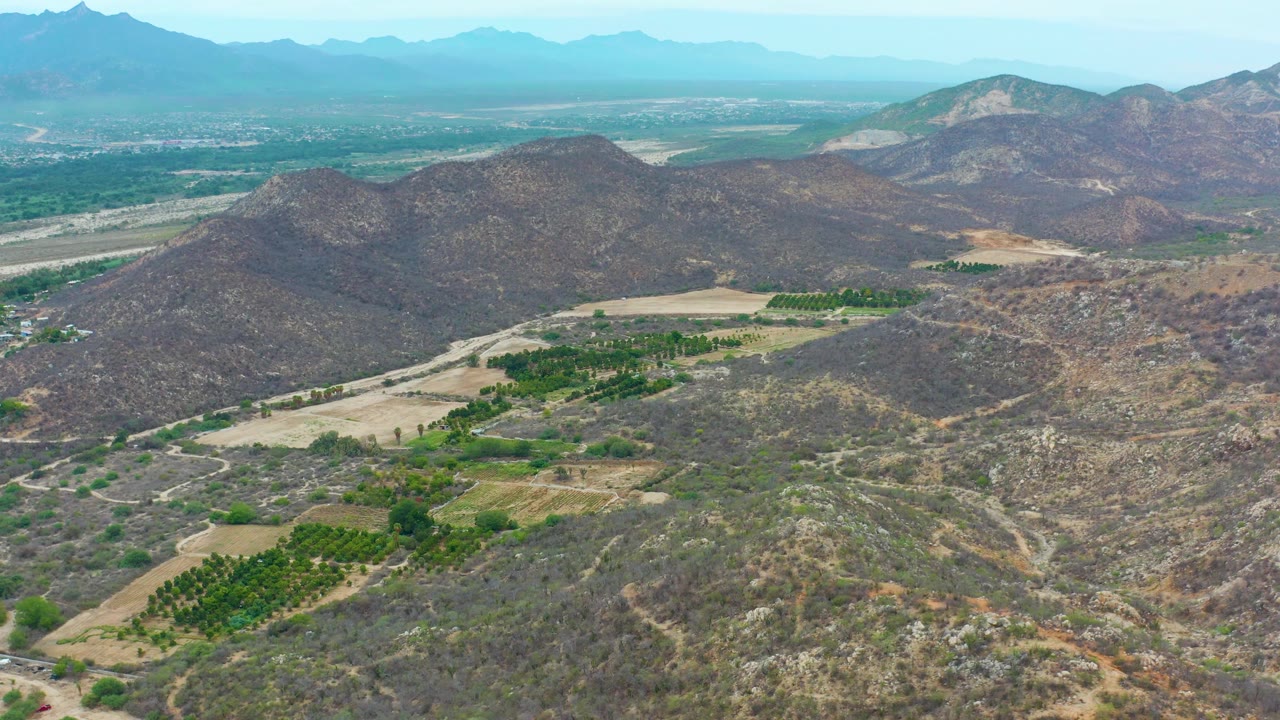 Wide La Playita Los Cabos aerial frame with dry brushy slopes in the foreground, green cultivated rows on the lower left and center, hazy background mountains, and no visible people.