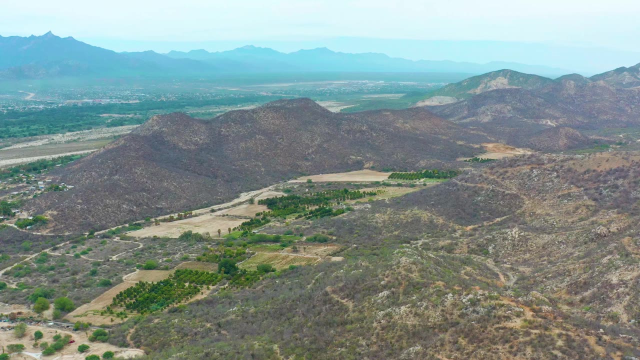 Aerial view in La Playita Los Cabos, Mexico, looking across a central rocky ridge toward rectangular green fields, pale valley tracks, blue haze, and no visible people.