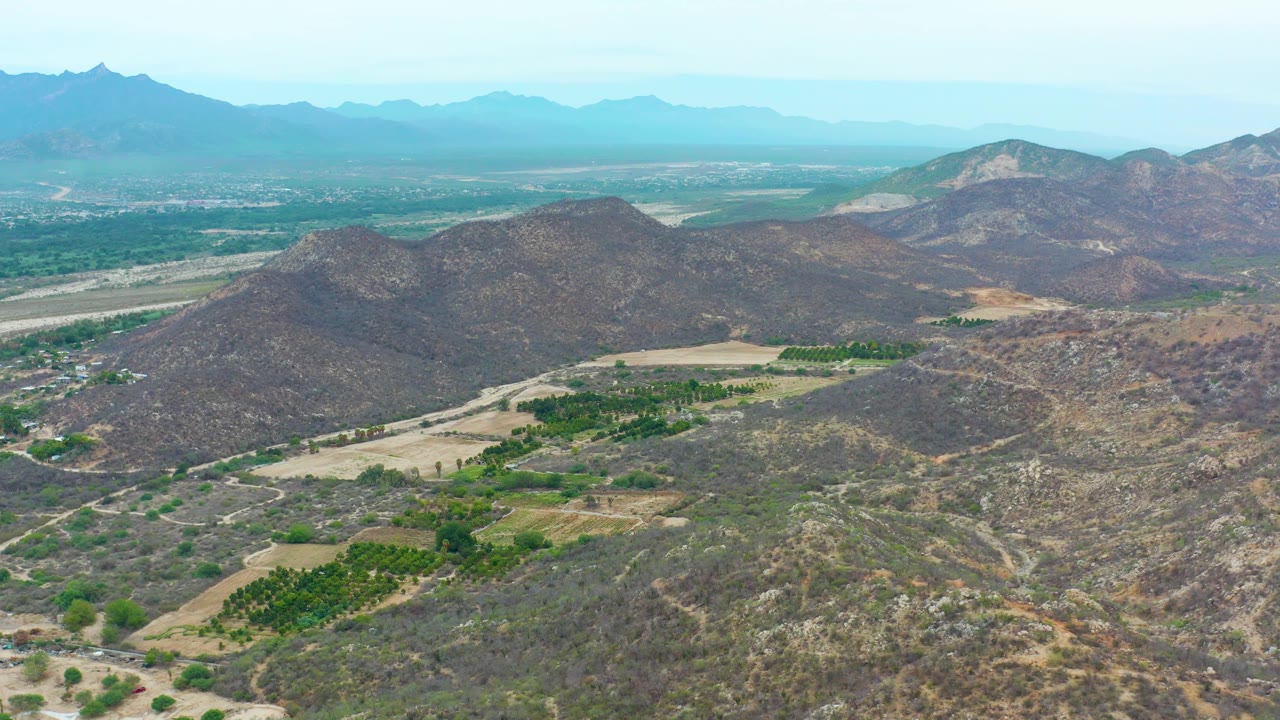 La Playita Los Cabos aerial frame showing dry scrub-covered hills, small green farm plots, a broad valley floor, distant blue mountains, and no visible people.
