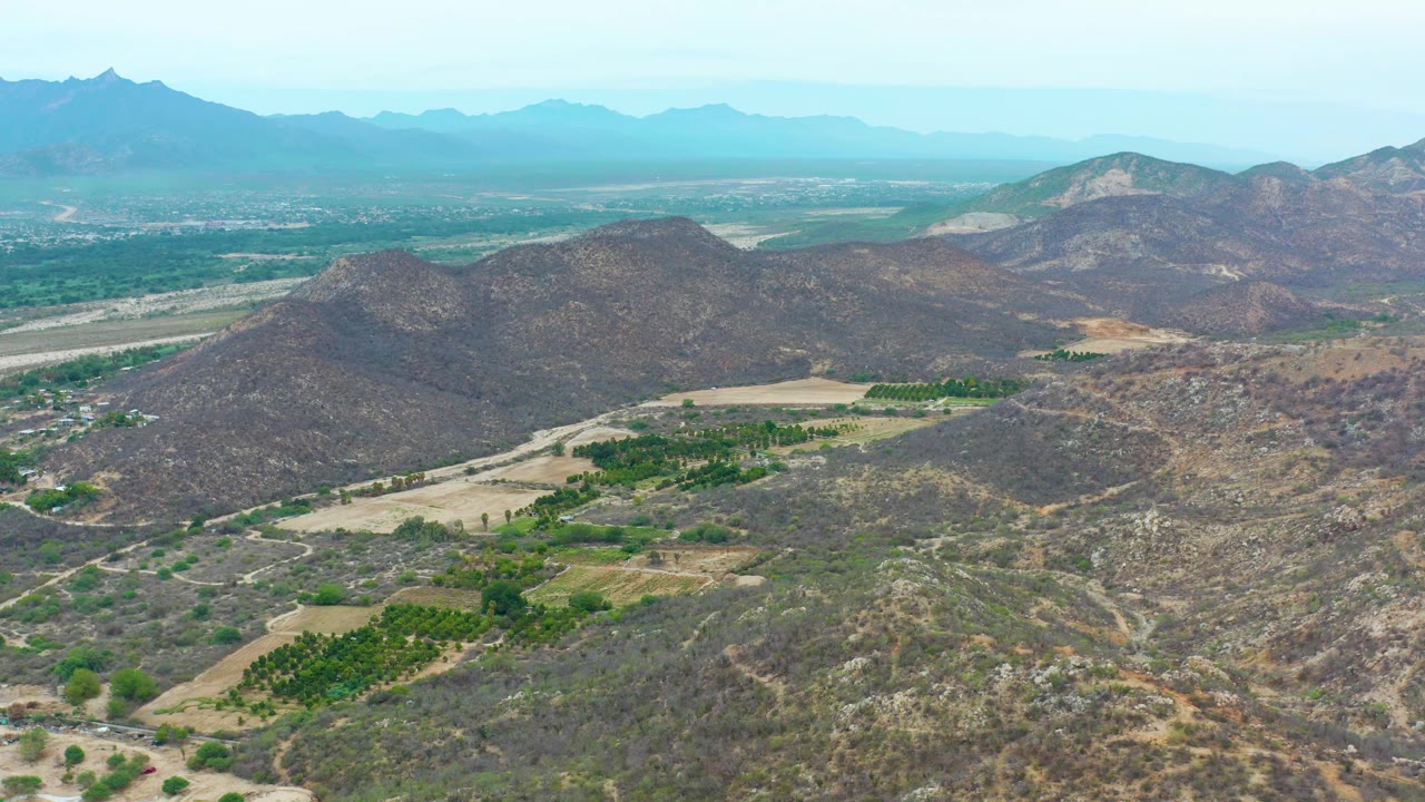 Mid-clip aerial view over La Playita Los Cabos, Mexico, with the green valley patches shifted in frame beneath rocky hills, layered mountains in back, and no visible people.