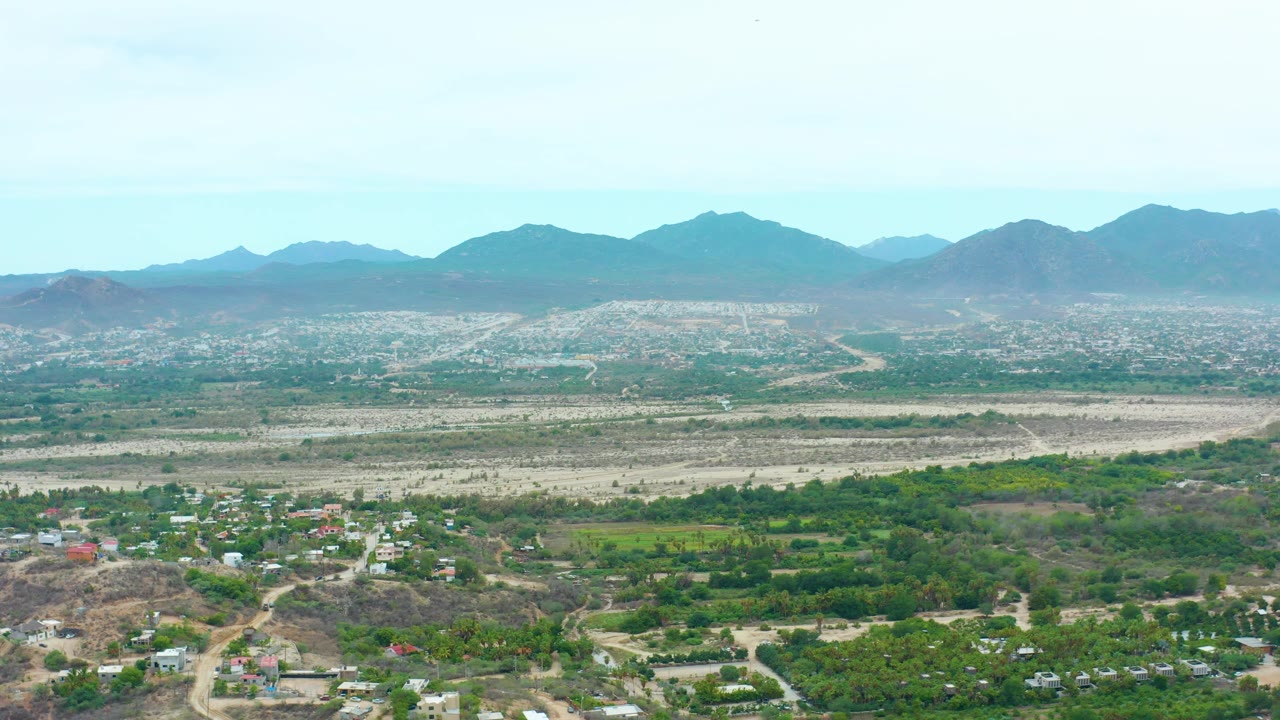 Opening frame of La Playita Los Cabos showing scattered homes and dirt roads below, green vegetation across the valley, dry washes, distant mountain ridges, pale sky, and no visible people.