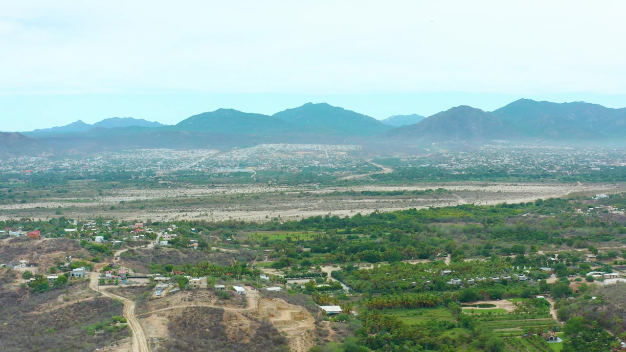 Wide aerial frame over La Playita Los Cabos showing green vegetation, scattered buildings, pale roads, a dry plain, distant mountain ridges, and no visible people.