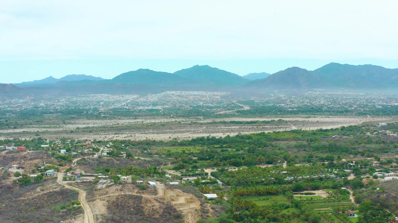 Aerial view in La Playita Los Cabos with foreground hills, green plots, small buildings, a pale riverbed-like plain, distant mountains, and no visible people.