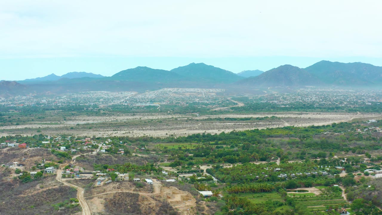 High view over La Playita Los Cabos showing rural greenery, light-colored dirt roads, scattered structures, a broad dry plain, blue-gray peaks, and no visible people.