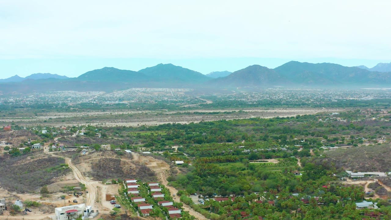 Opening aerial frame over La Playita Los Cabos showing a green valley, dusty hillside roads, scattered buildings, distant mountain ridges, hazy sky, and no visible people.