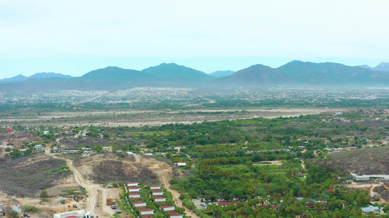 La Playita Los Cabos aerial frame with hillside construction areas in the lower frame, a broad green valley through the center, pale mountain ranges beyond, and no visible people.