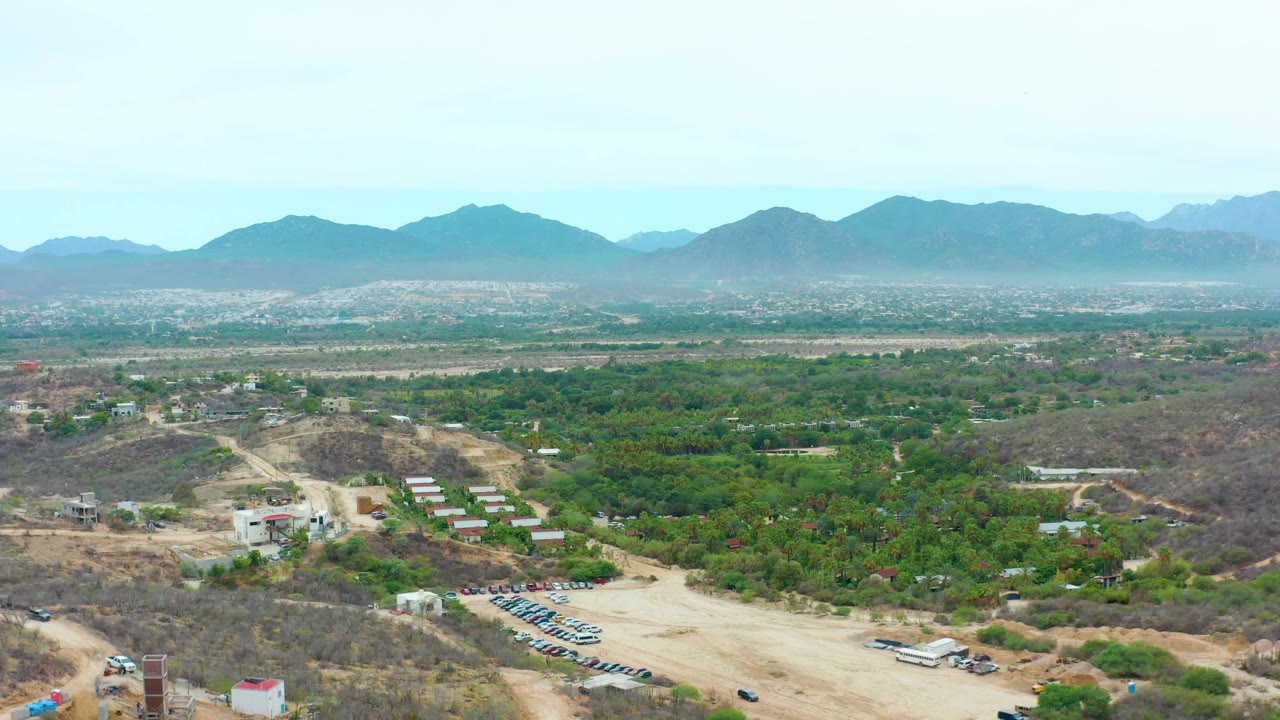 Aerial frame over La Playita Los Cabos showing a pale dirt road, green tree cover, scattered buildings, parked vehicles, dry hills, hazy mountains, and no visible people.
