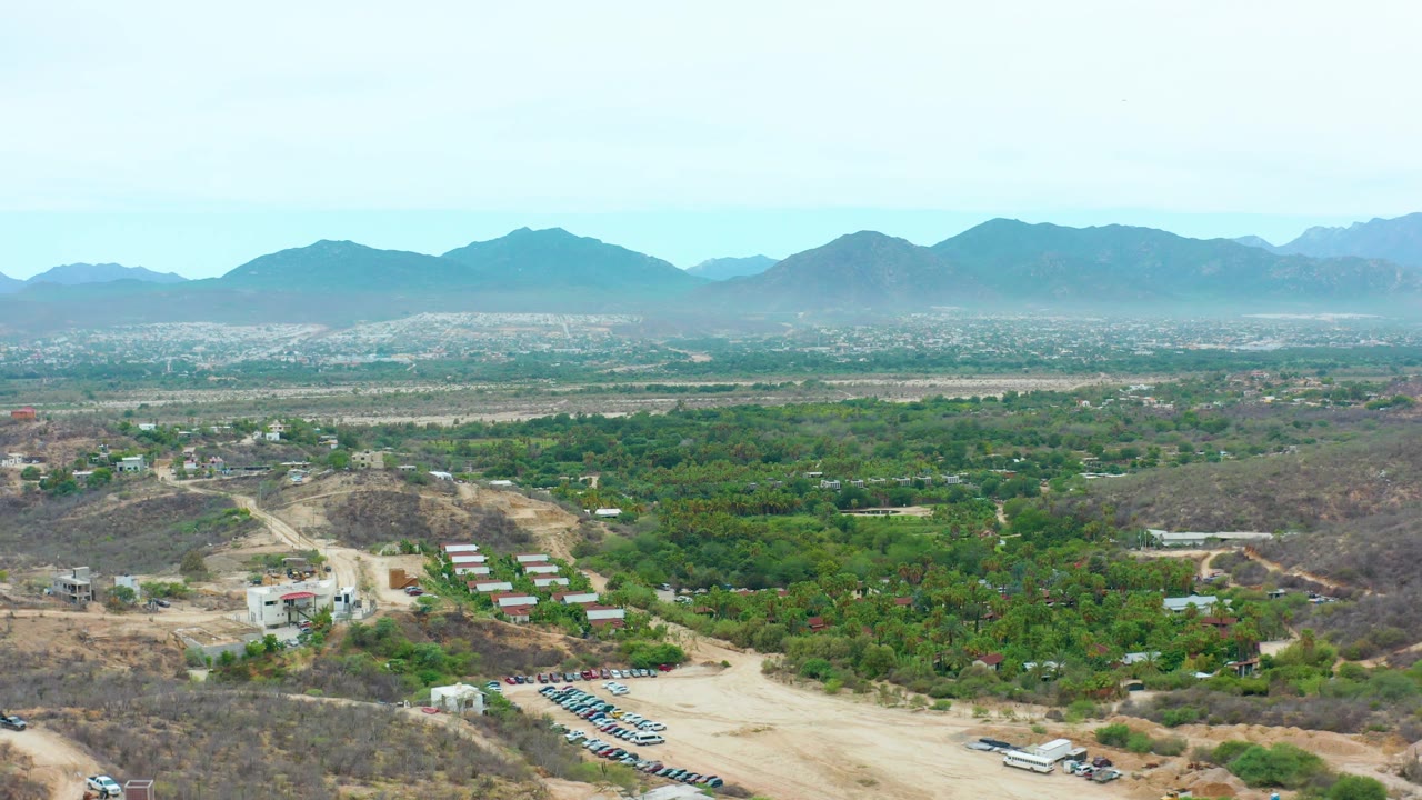 La Playita Los Cabos aerial frame with a light dirt clearing in the foreground, small structures along the slope, broad green tree cover, hazy mountain ridges, and no visible people.