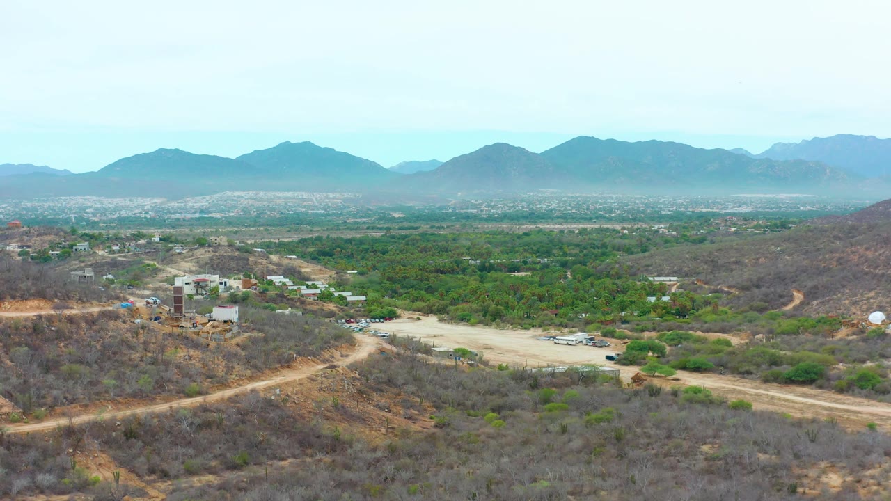 Opening aerial frame over La Playita Los Cabos shows dry scrub hills, pale dirt roads, scattered buildings, a green valley, and hazy mountains with no visible people.