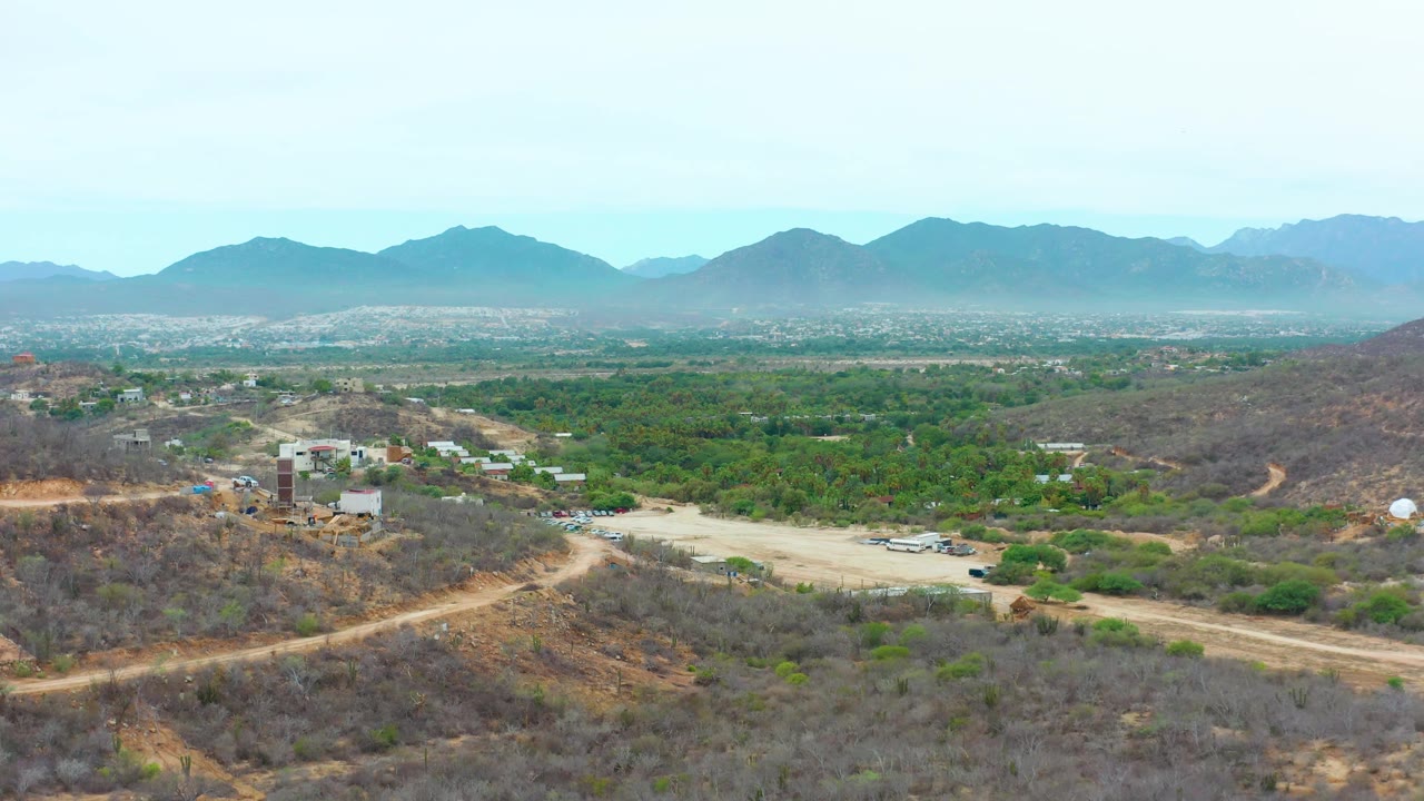 At 0:01 the La Playita Los Cabos aerial view keeps the curving dirt road and hillside buildings low in frame, with the green valley and mountain range stretching behind and no visible people.