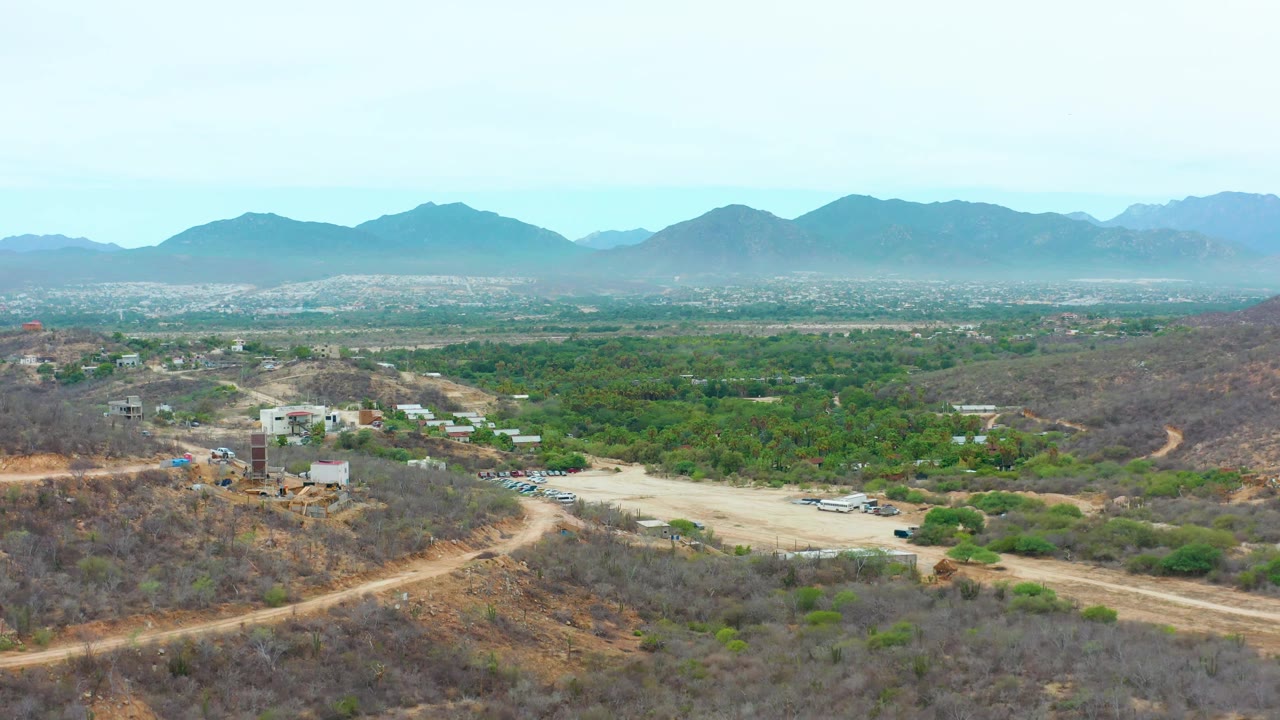 At 0:02 the frame shows the dry hillside and parking area slightly closer, with small structures, parked vehicles, green vegetation across the valley, and distant mountains in La Playita Los Cabos, with no visible people.