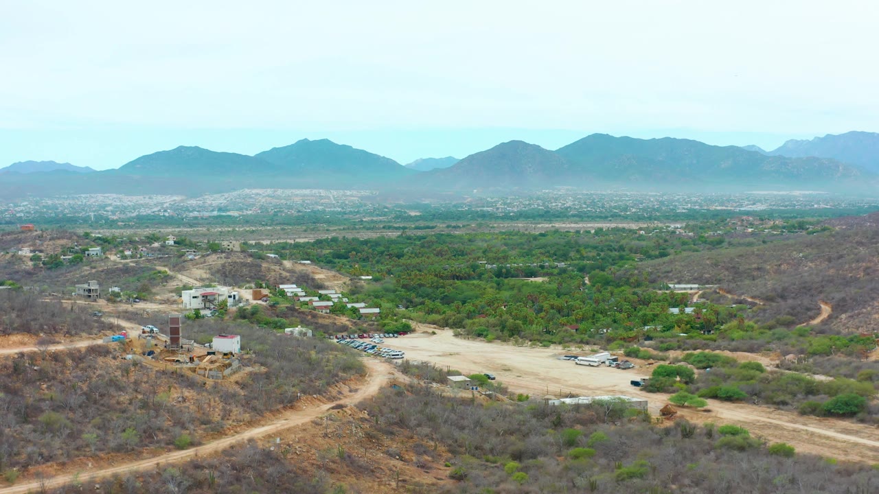 At 0:03 the aerial frame over La Playita Los Cabos balances dry foreground hills and dirt tracks with a broad green valley and hazy blue mountain backdrop, with no visible people.