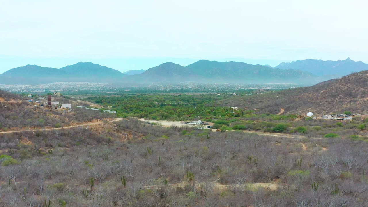 Opening frame of La Playita Los Cabos from above, with dry scrubby foreground hills, a green valley, scattered buildings, distant blue mountains, pale sky, and no visible people.