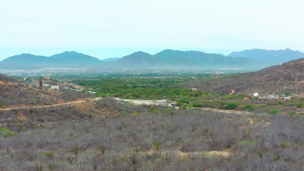 Aerial landscape with scrub hills, green vegetation, a dirt road, buildings, and mountain ridges.