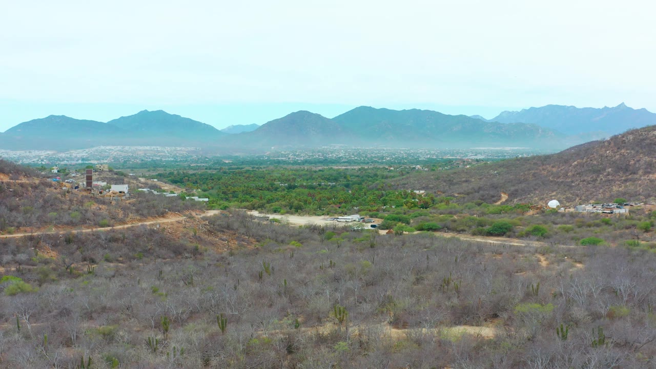 La Playita Los Cabos aerial frame with arid brushland and cacti in the foreground, a road tracing the hillside, low buildings near the valley, layered mountains, and no visible people.