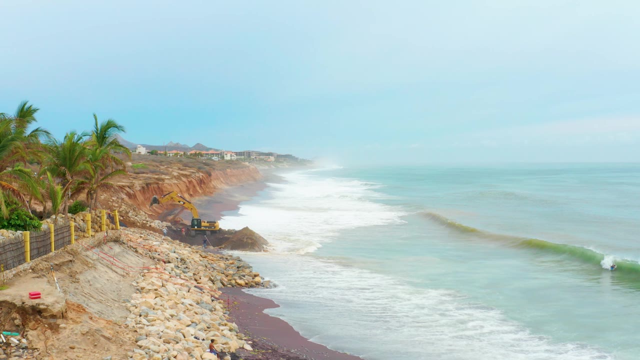 Wide view of La Playita Los Cabos shoreline with palm trees on the left, rocks below the cliff, white surf rolling in, a yellow construction vehicle near the beach, and no visible people.
