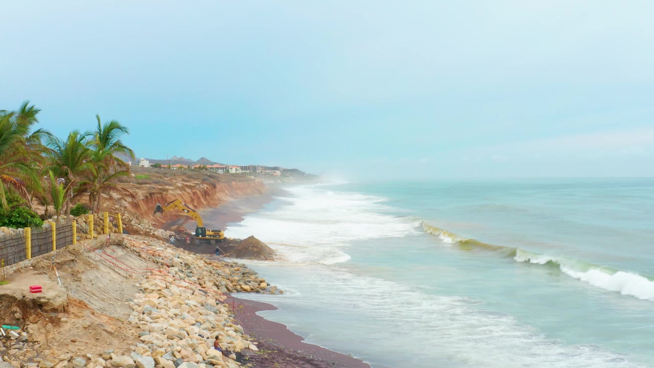 Elevated La Playita Los Cabos shoreline view with a larger white wave breaking near the right side, palm trees and a cliff fence at left, rocks below, and no visible people.