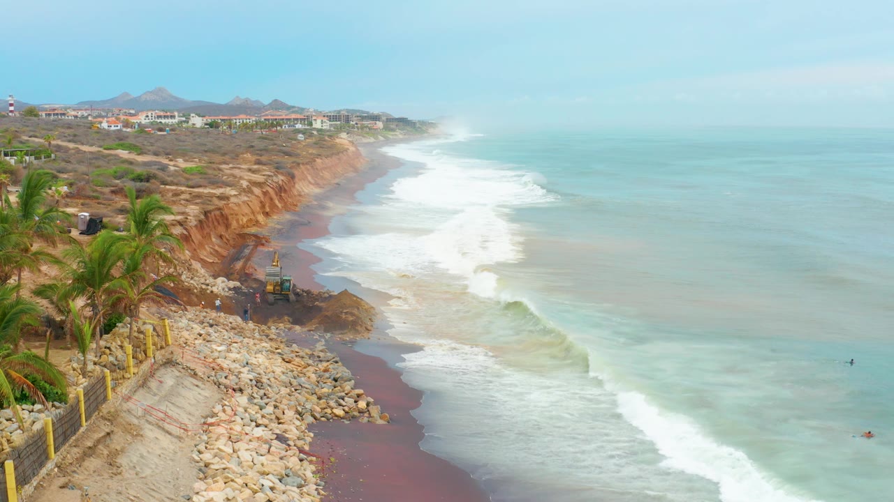 At La Playita Los Cabos, the excavator remains near the cliff face as white waves roll along the narrow beach, with palms and a roadside fence in the foreground and no visible people.