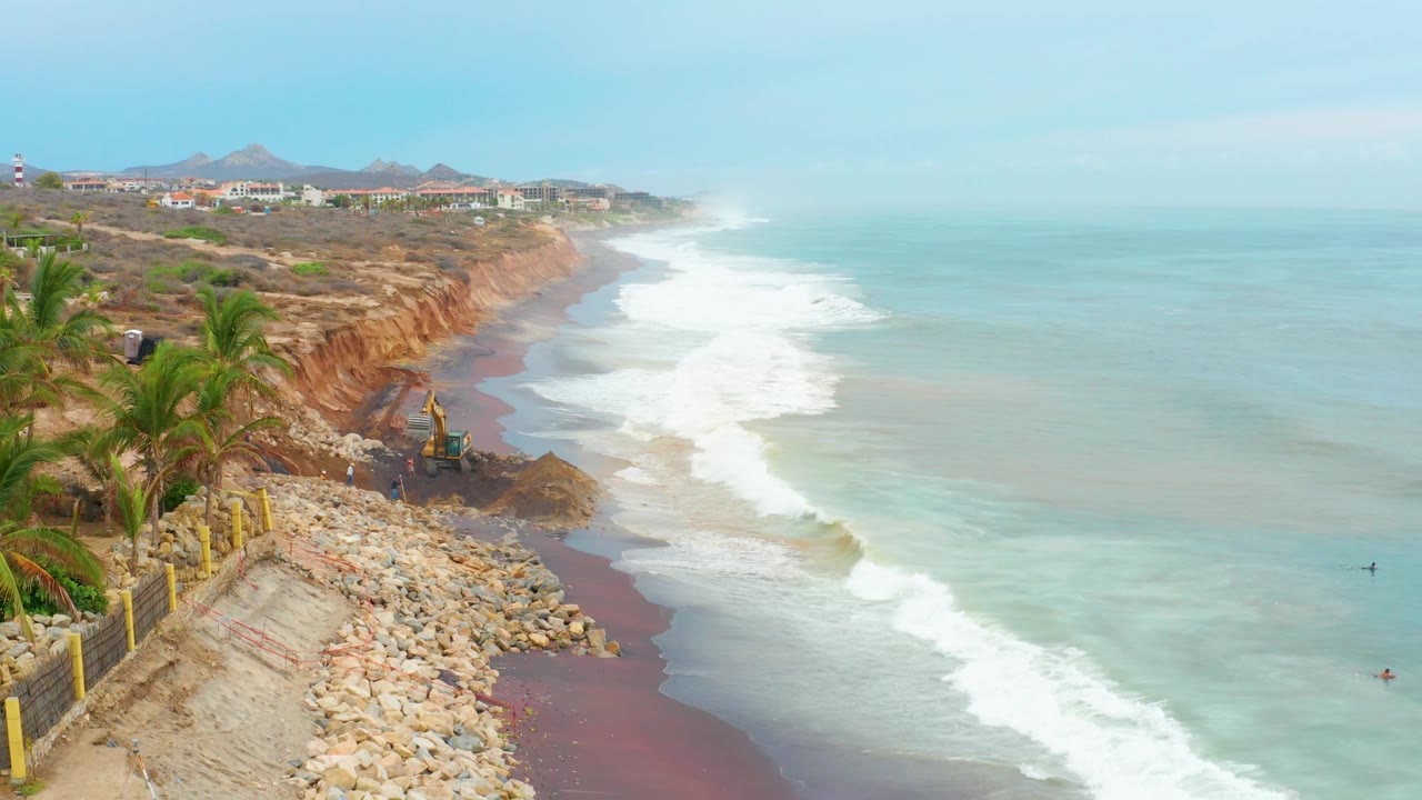 At La Playita Los Cabos, the rocky slope and excavator fill the left side while broad pale surf and blue ocean stretch rightward toward distant buildings, with no visible people.
