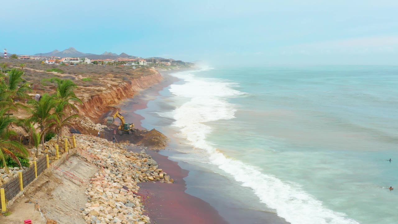 At La Playita Los Cabos, the excavator works near a darker patch of cliff material above red-brown sand and foamy waves, with palms and a fence at left and no visible people.