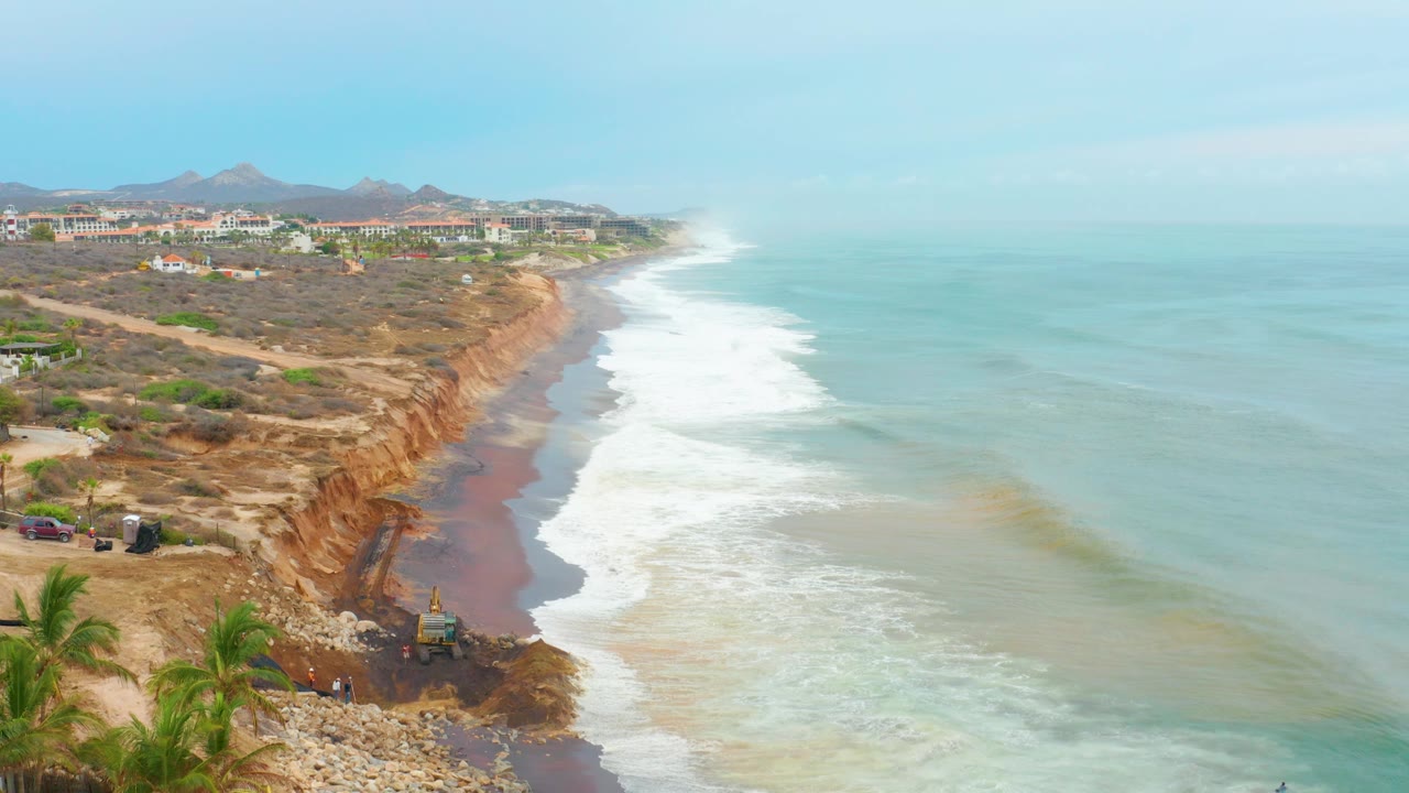 Opening frame of La Playita Los Cabos shoreline with white surf running along orange-brown cliffs, palm fronds at lower left, rocky riprap in front, and coastal buildings in the distance.