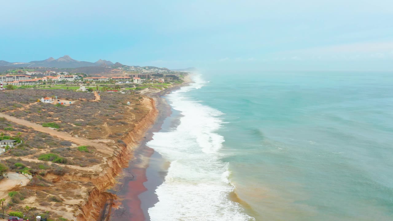 Opening aerial frame over La Playita Los Cabos, Mexico, showing eroded brown cliffs at left, white waves breaking along the shore at right, distant buildings inland, and no clearly visible people.
