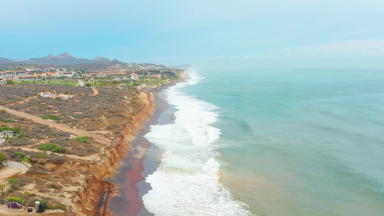 Aerial frame over La Playita Los Cabos with dry cliff terrain, a dirt road near the edge, tiny vehicles near the lower left, white surf along the beach, and distant developed buildings behind the cliffs.