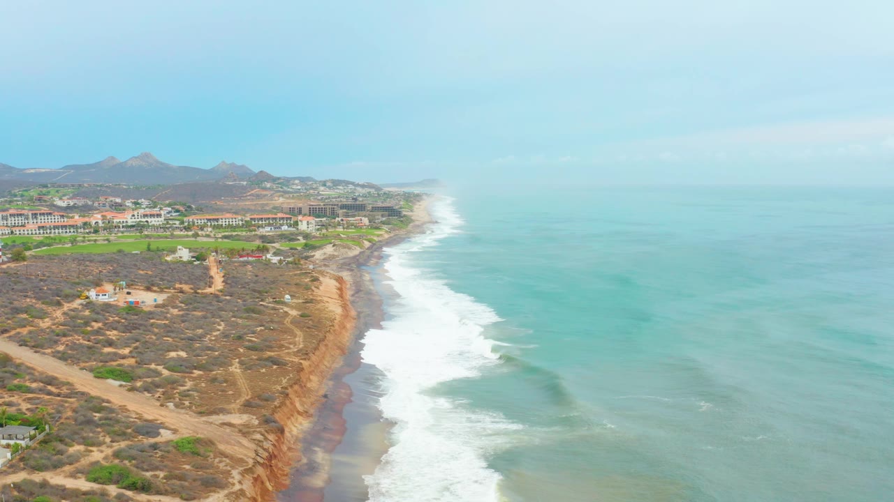 Aerial frame over La Playita Los Cabos showing a longer line of white waves beside the cliff base, arid ground and dirt paths on the bluff, low buildings in the distance, and no visible people.