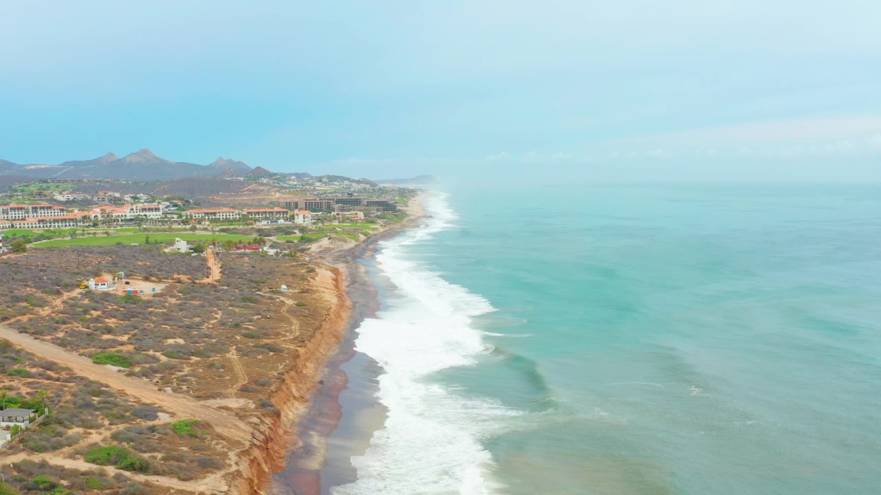 Aerial view of La Playita Los Cabos with the ocean occupying most of the right side, foamy waves approaching shore, reddish-tan cliff faces, dry dunes, distant buildings, and no visible people.