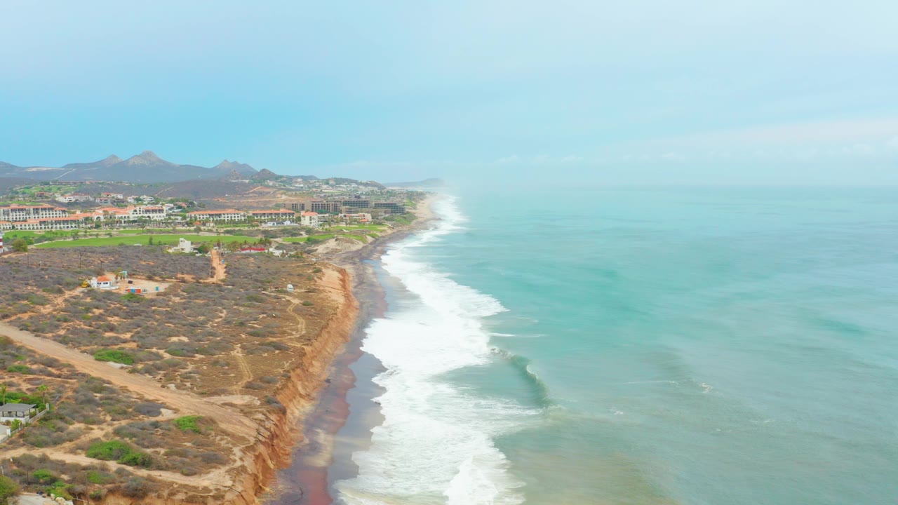 Mid-clip aerial frame over La Playita Los Cabos showing rolling surf curving along the beach, scrubby dry bluff terrain, a dirt road or track, low buildings inland, and no visible people.