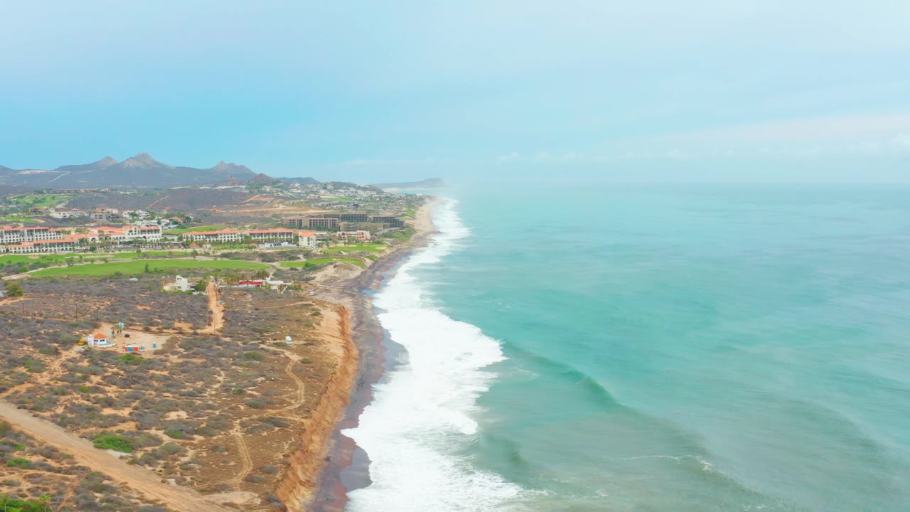 At 0:00, an aerial view over La Playita Los Cabos shows turquoise ocean, white surf, dry hillside tracks, low inland buildings, distant hills, and no visible people.