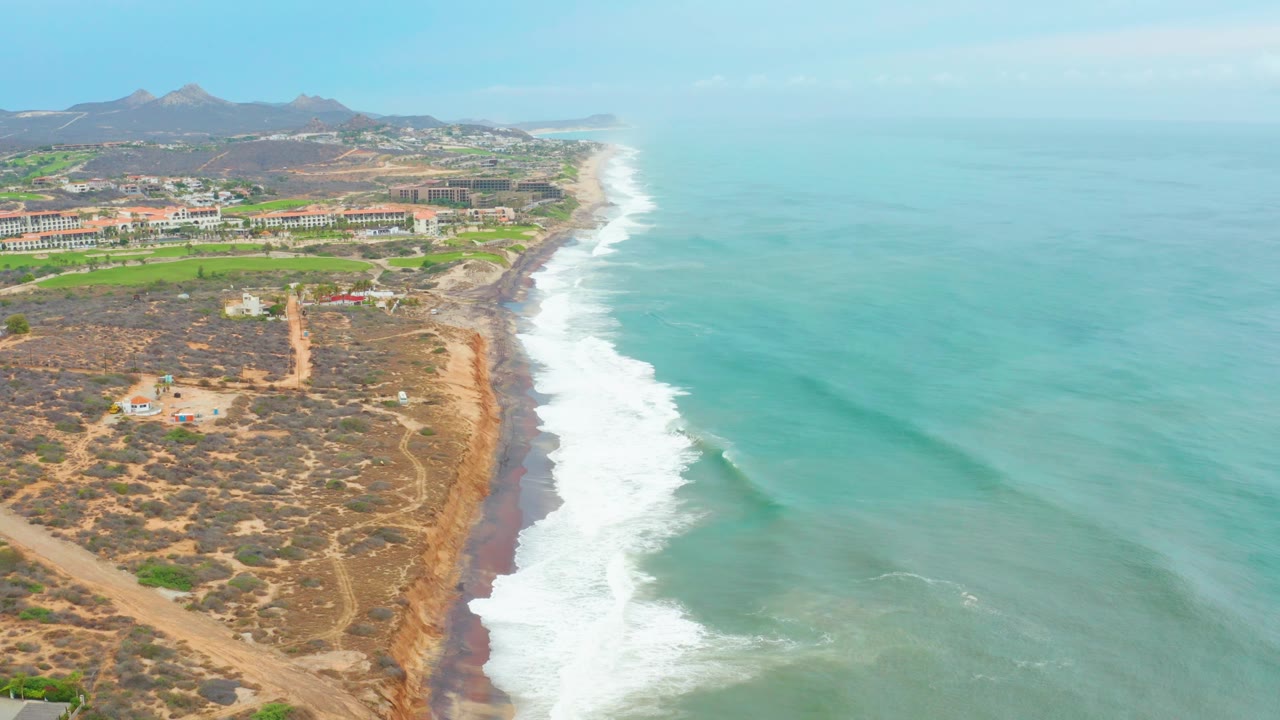 Drone aerial over La Playita Los Cabos coastline with a wider band of white surf near center, dry coastal scrub and paths at left, low buildings beyond, and no visible people.