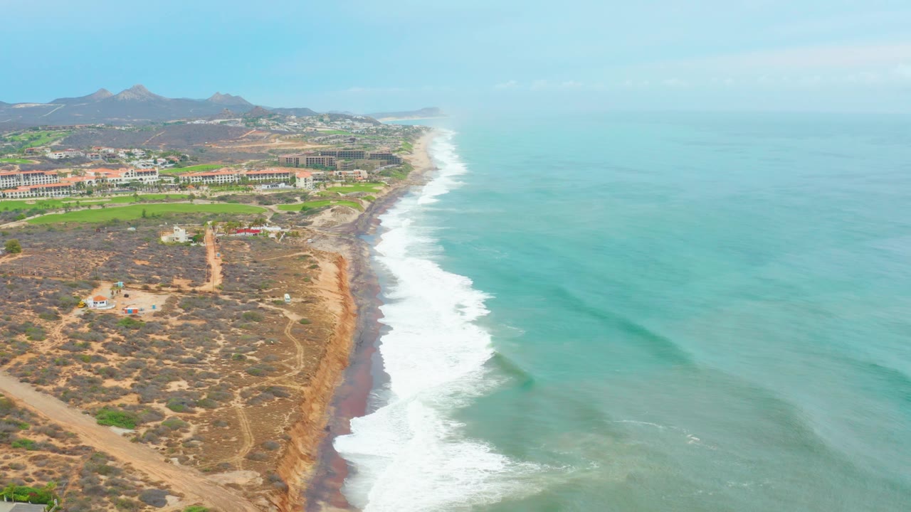 La Playita Los Cabos aerial view with white breakers running up the shore, arid land and a dirt road at left, open turquoise ocean at right, hazy sky, and no visible people.