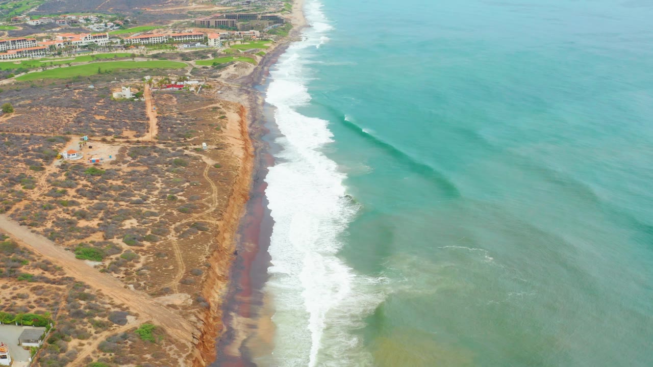 Aerial frame at 0:01 shows La Playita Los Cabos waves curling closer to shore, with scrub-covered dunes and dirt paths left of the foamy beach and no visible people.