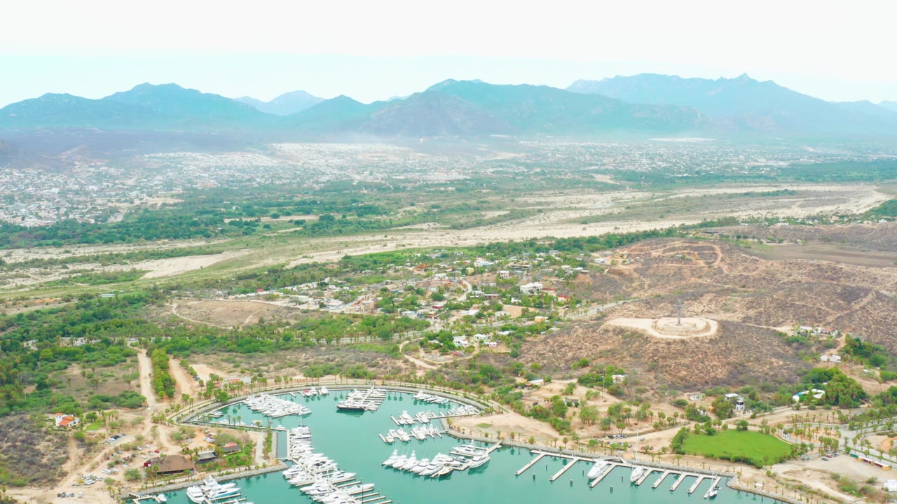Drone frame over La Playita Los Cabos showing boat docks in teal water at the lower left, hillside paths and scattered buildings across dry terrain, a runway and city in the distance, and no visible people.