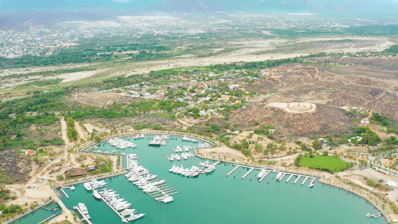Opening aerial frame over La Playita Los Cabos, Mexico, showing turquoise marina water, white docked boats, shore roads, green vegetation, dry hills, hazy mountains, and no visible people.