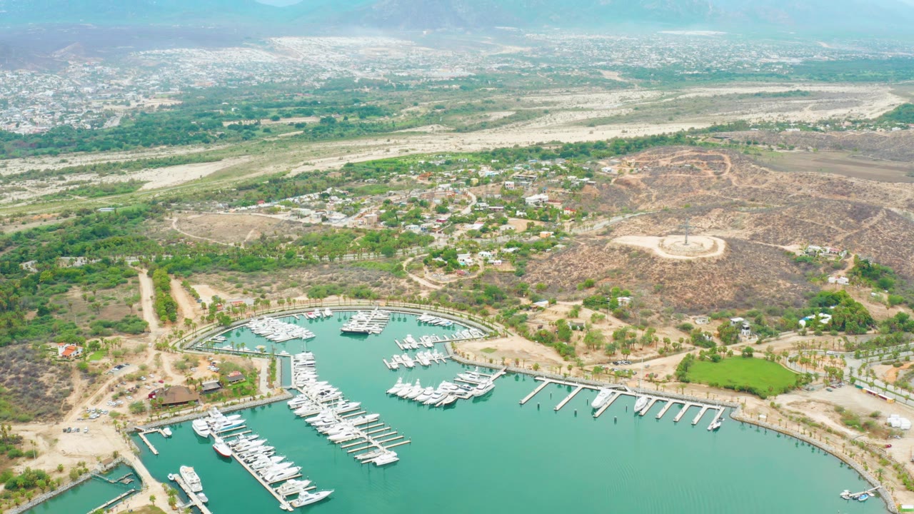 Early aerial frame of La Playita Los Cabos marina with clustered white boats in turquoise water, docks along the shore, scattered buildings, dry hills, distant mountains, and no visible people.