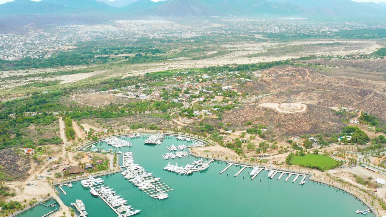 Aerial frame over La Playita Los Cabos with marina docks and white boats across the lower frame, green vegetation and buildings in the middle distance, dry hills, hazy mountains, and no visible people.