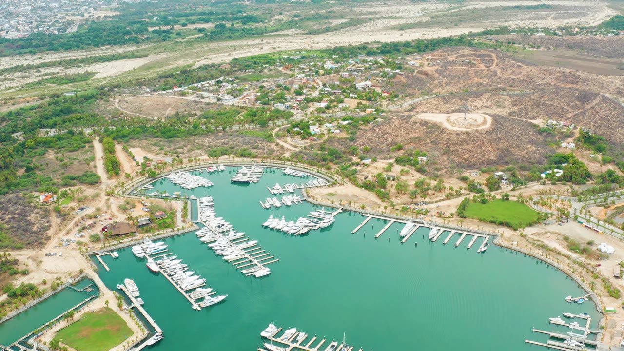 Aerial view at 0:01 over La Playita Los Cabos showing the curved marina basin, rows of docked boats, pale roads, scrubby hills, and no visible people.