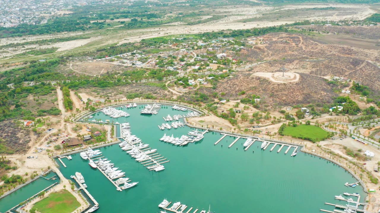 Frame 0:02 shows La Playita Los Cabos marina from above, with docked boats clustered near the curved shoreline, hillside roads behind, and no visible people.
