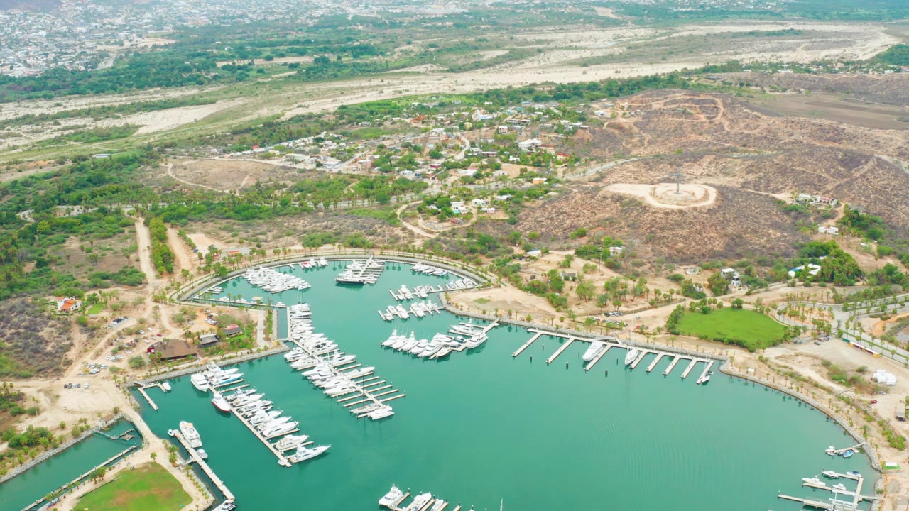 Mid-clip aerial frame over La Playita Los Cabos with marina slips at the bottom, a dry hill and green areas in the center, distant buildings above, and no visible people.