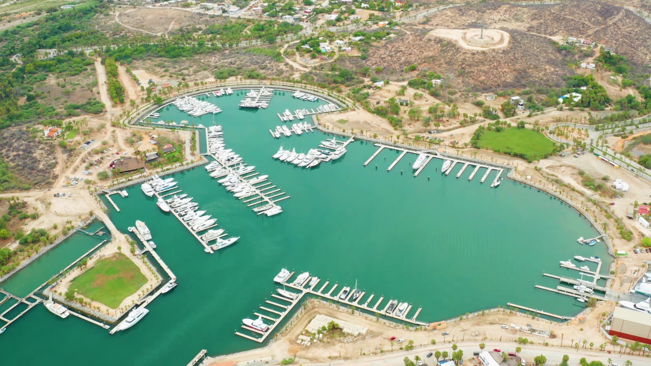 Aerial view over La Playita Los Cabos marina with turquoise water, white docked boats, shoreline roads, dry hills, and no visible people at the opening moment.