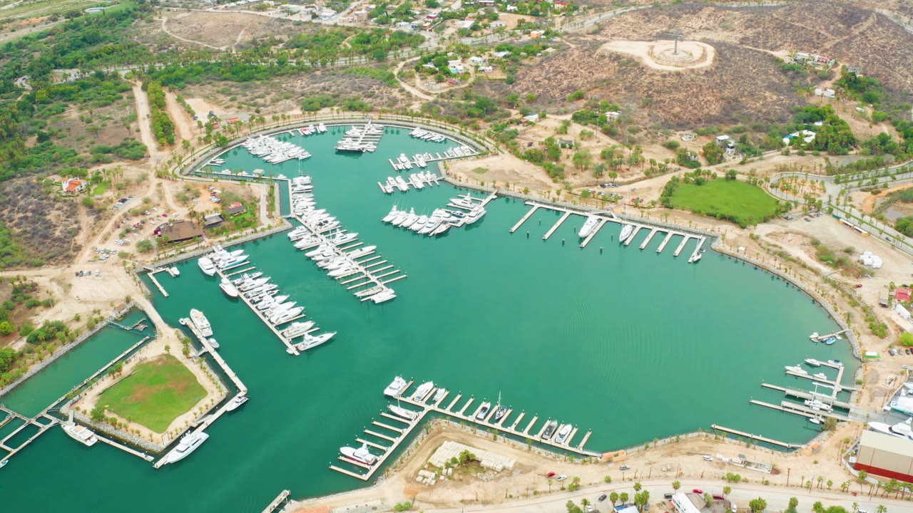 High aerial frame of La Playita Los Cabos marina showing clustered boats along long white slips, green-blue water, dusty shore roads, hillside vegetation, and no visible people.